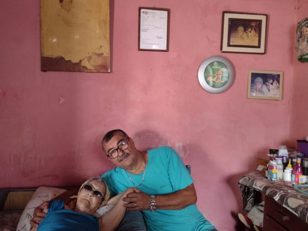 Nair e Paulo, com fotos de Fatinha ao redor da parede. Este cômodo, no passado, era sala de dia e cama de noite. Havia uma beliche e a cama de Fatinha - (Foto: João Ramos/Midiamax)